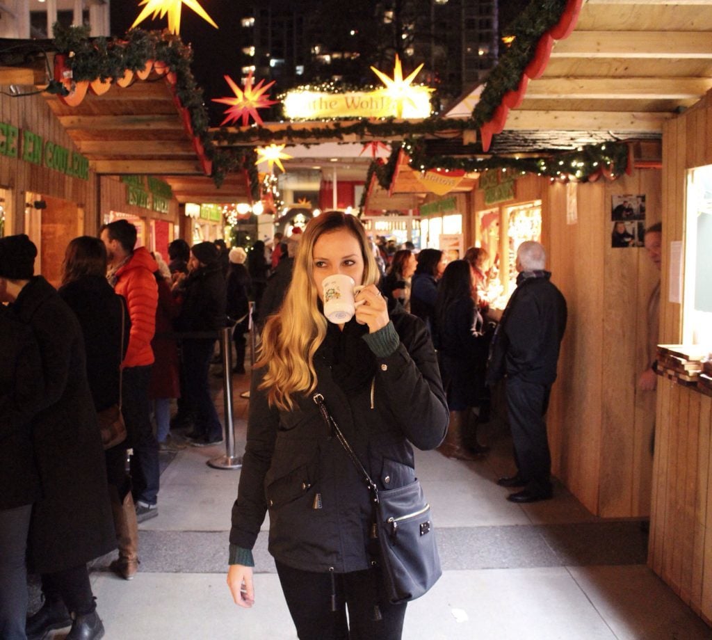 Maddy drinking from a white mug while wandering around the Vancouver Christmas Market crowded with people