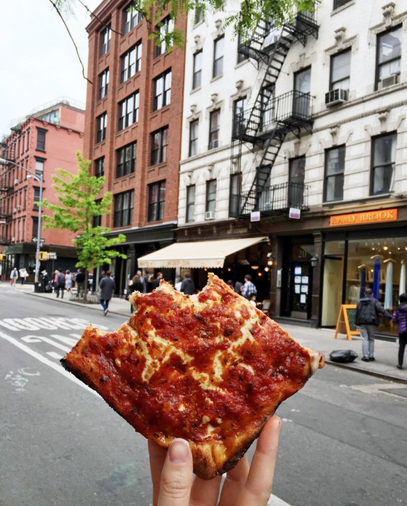 Holding up a square slice of pizza in front of some shops in Manhattan