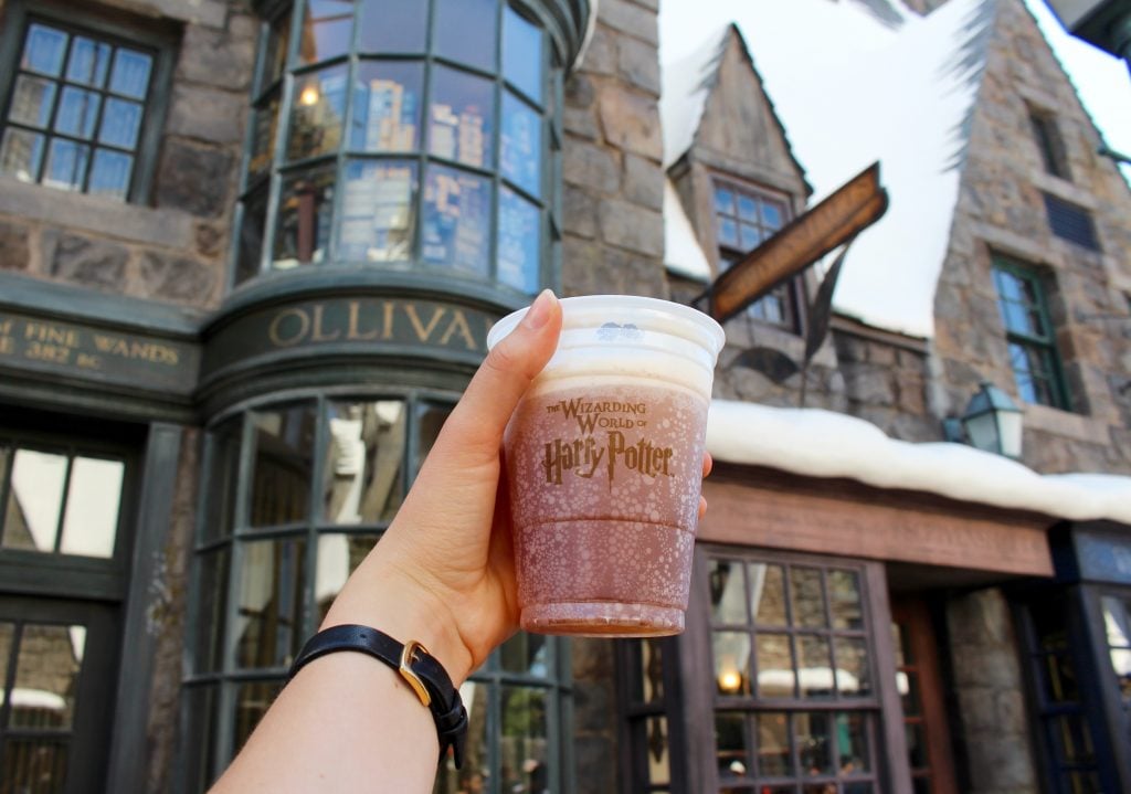 Holding up a cold drink in a cup in front of the Ollivanders. Consider the do's and don'ts of visiting The Wizarding World of Harry Potter in this guide for a more fun trip.