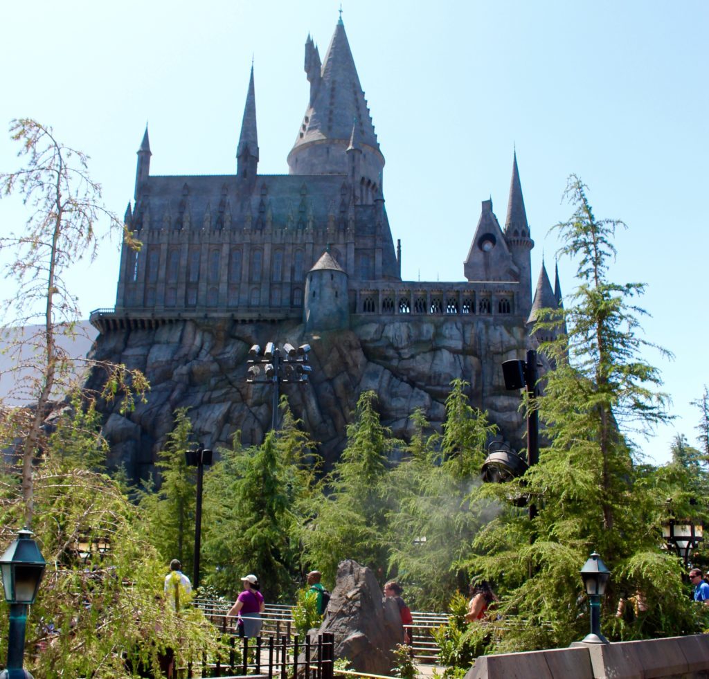 Hogwarts castle on top of a rocky mountain while tourists walk around along the walkway surrounded by pine trees