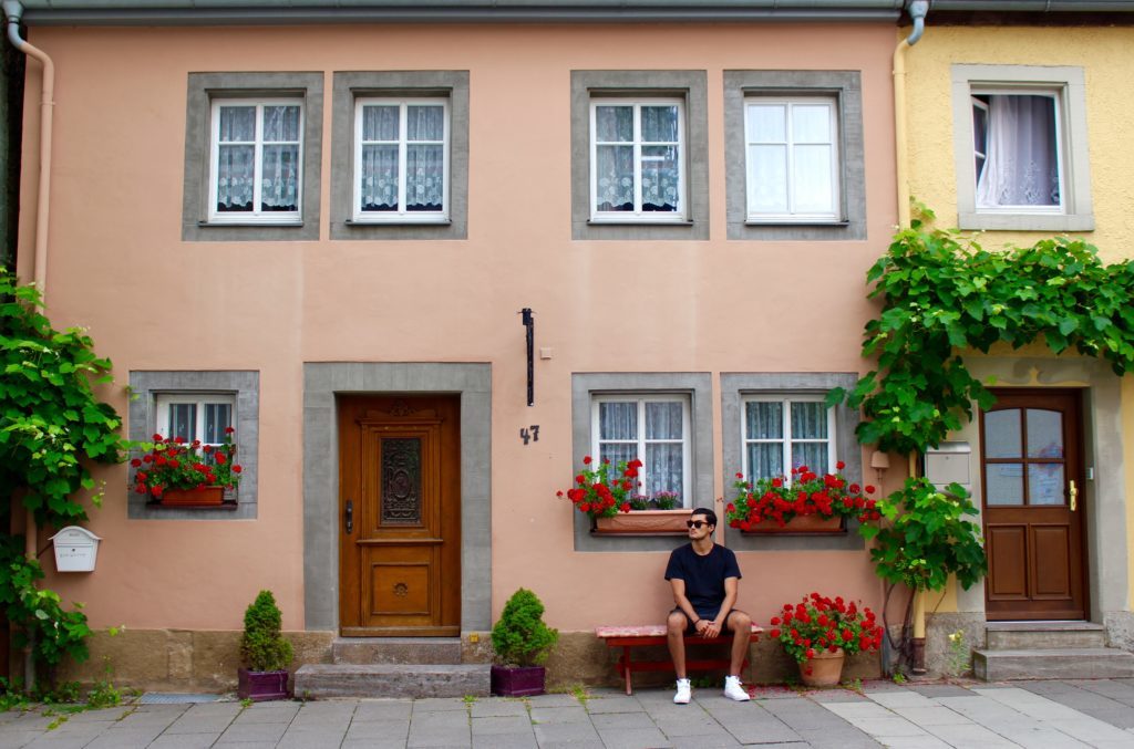 Mauricio, blogger, sitting on a chair outside a house decorated with potted flowers. The old town of Rothenburg ob der Tauber is one of the best places to visit in Germany.