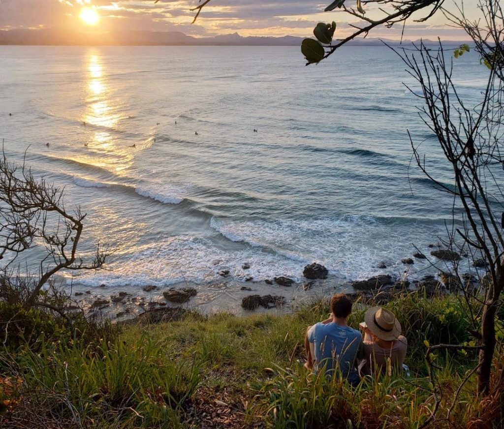 Maddy and her friend sitting on grass by the beach while admiring the beautiful sunset. Before traveling with a friend, be sure to ask the right questions.