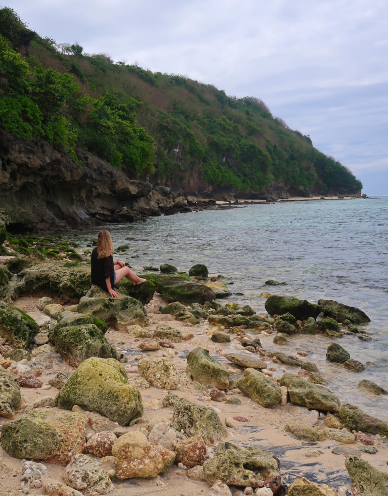 Maddy, blogger, sitting on mossy rocks by the beach. Relaxing on the beautiful beaches of Jimbaran is the perfect way to end your three weeks in Bali.