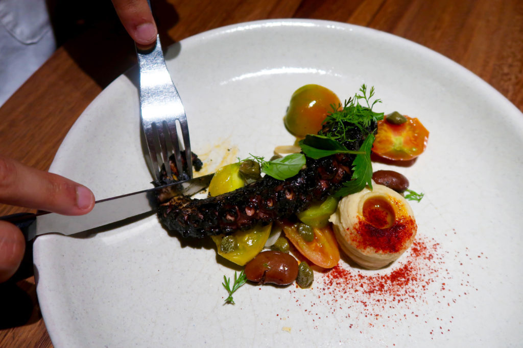 Cutting a cooked octopus tentacle garnished with spices, herbs, and tomatoes