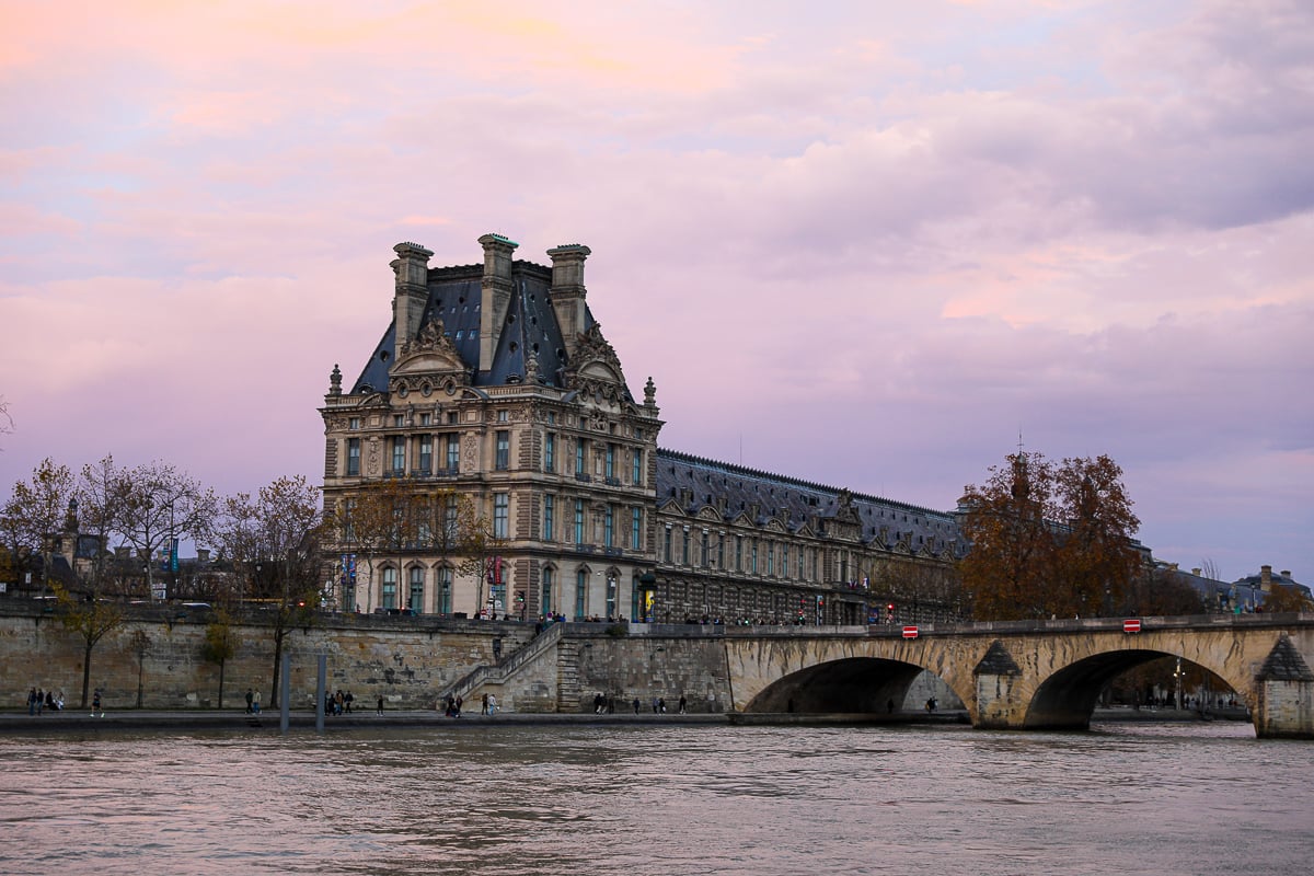 View of the Louvre Museum along the Seine River at sunset in Paris, with the Pont Royal bridge and soft pink and purple clouds in the sky.