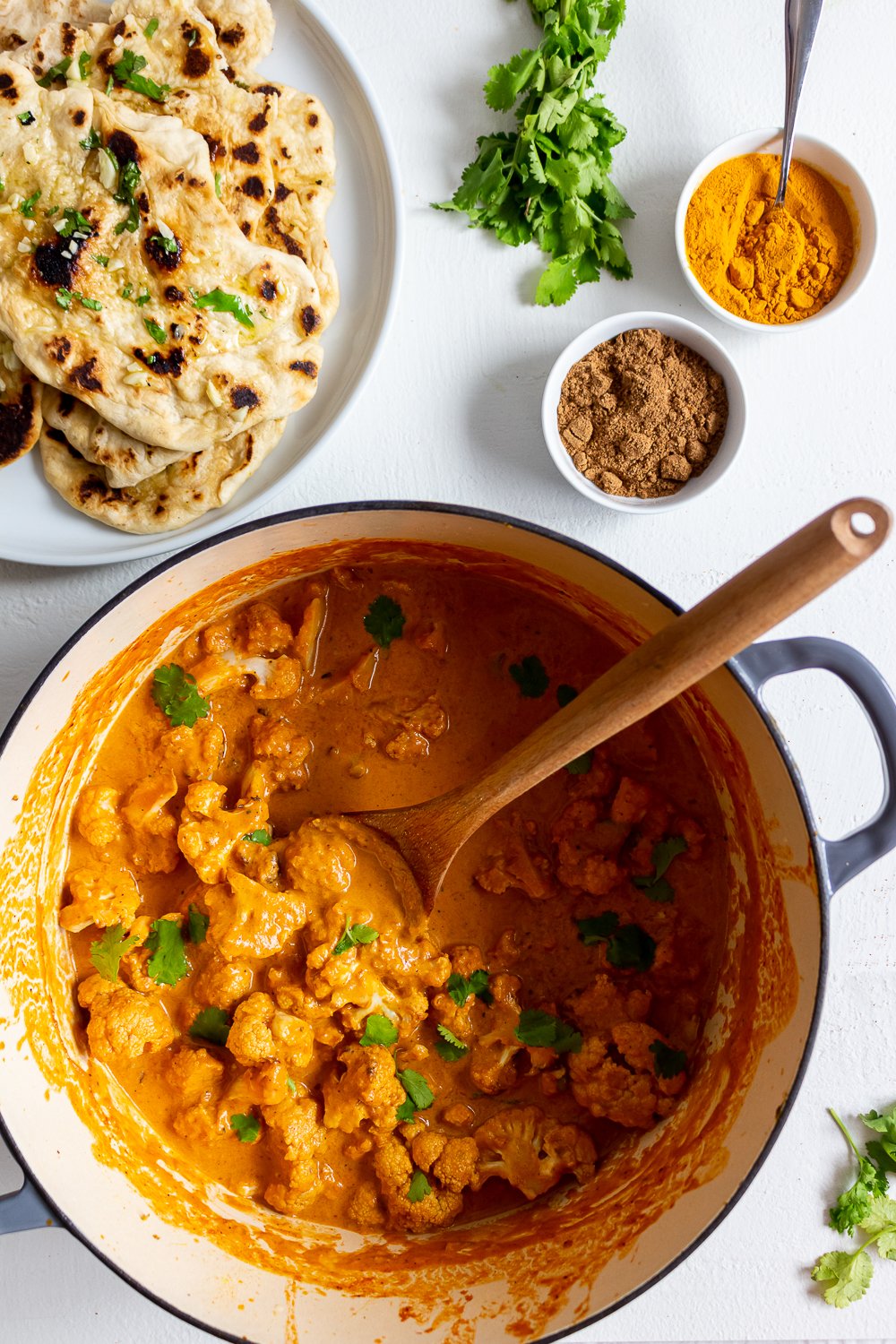 A pot of cauliflower tikka masala sprinkled with cilantro and a plate of garlic naan in the background