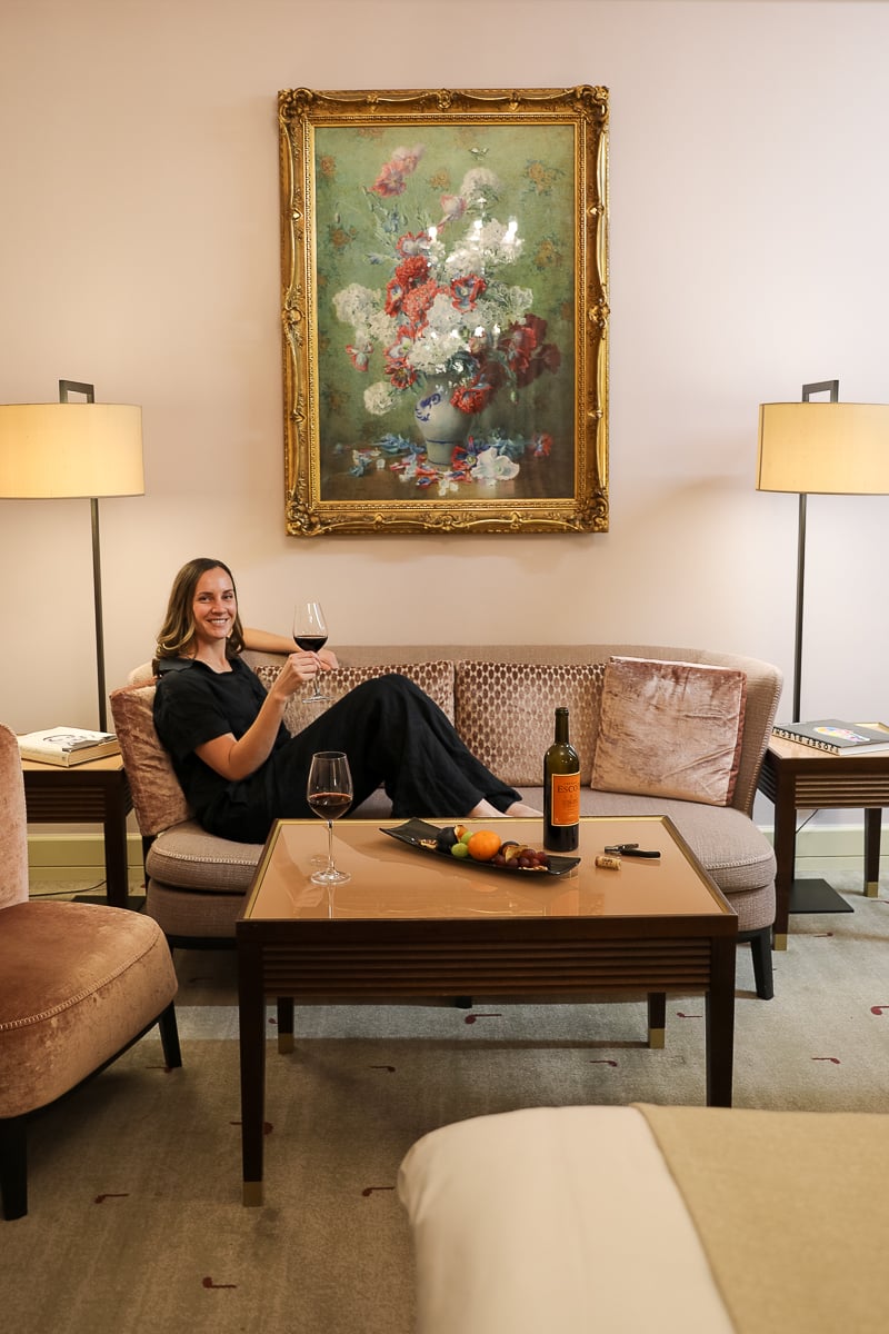 Maddy relaxing with a glass of red wine in a luxury suite at Aria Hotel Prague, featuring elegant décor, velvet sofa, artwork, and a bottle of wine on the table.