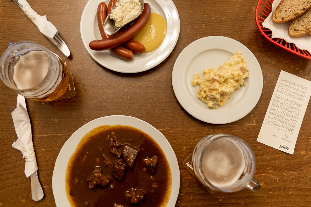 Traditional Czech meal at Lokál restaurant in Prague – beef goulash with bread dumplings, sausages with mustard and horseradish, potato salad, fresh bread basket, and two pints of Czech beer on a wooden table.