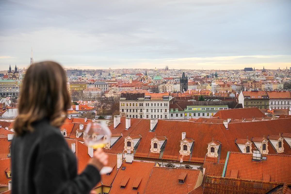 Maddy holding a glass of white wine while overlooking Prague’s red rooftops, historic churches, and city skyline at sunset.