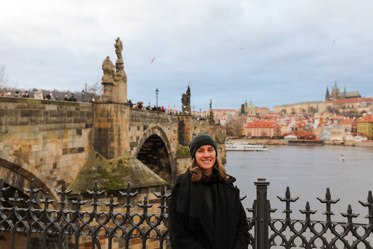 Maddy smiling in front of Prague’s Charles Bridge, with statues lining the historic stone bridge, the Vltava River flowing beneath, and Prague Castle and colorful rooftops in the background.