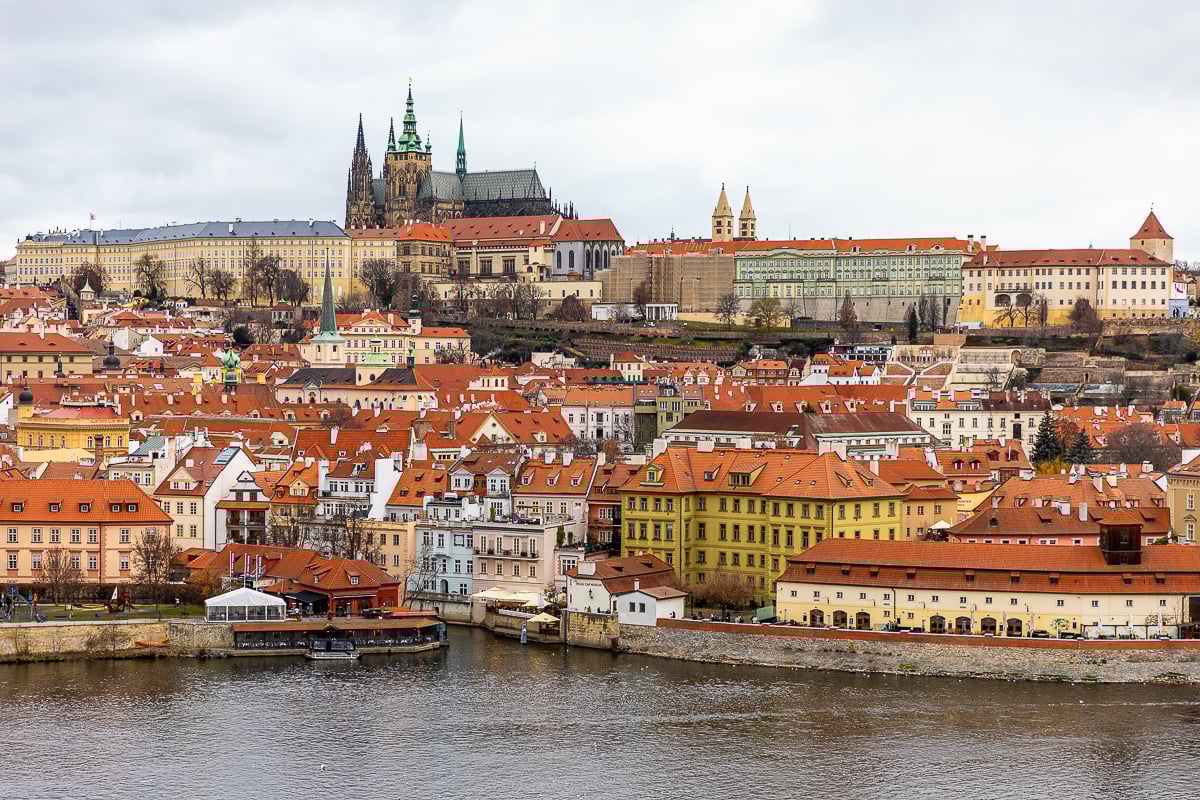 Panoramic view of Prague Castle complex and St. Vitus Cathedral rising above the red rooftops of Malá Strana, seen from across the Vltava River.