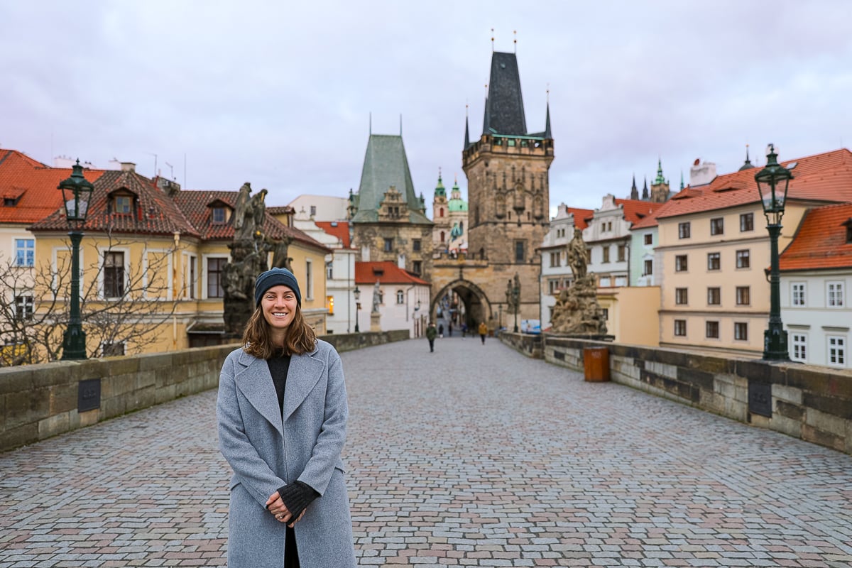 Maddy standing on Prague’s Charles Bridge on a quiet morning, with cobblestone walkway, statues, and the Gothic Old Town Bridge Tower in the background.
