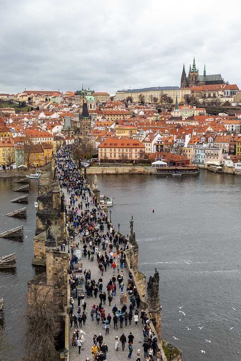 Crowds walking across Charles Bridge toward Malá Strana with Prague Castle in the background on a cloudy day.