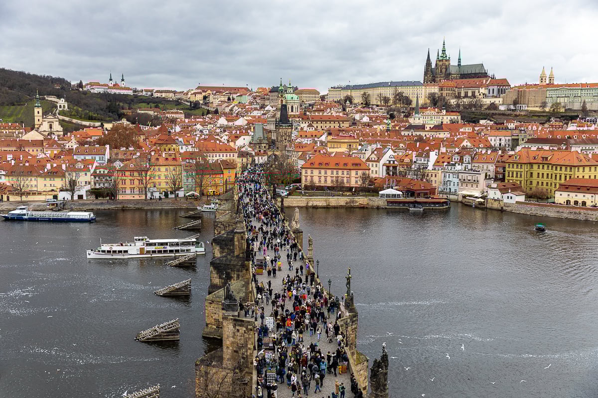View from the Old Town Bridge Tower overlooking Charles Bridge packed with people, the Vltava River, and Prague Castle rising above the red rooftops of Malá Strana.