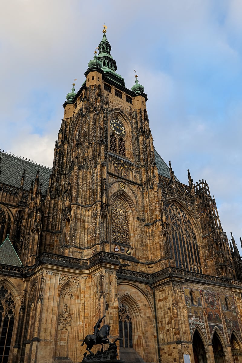 Tower of St. Vitus Cathedral at Prague Castle, showcasing Gothic spires, ornate stonework, and astronomical clock detail against a blue sky.