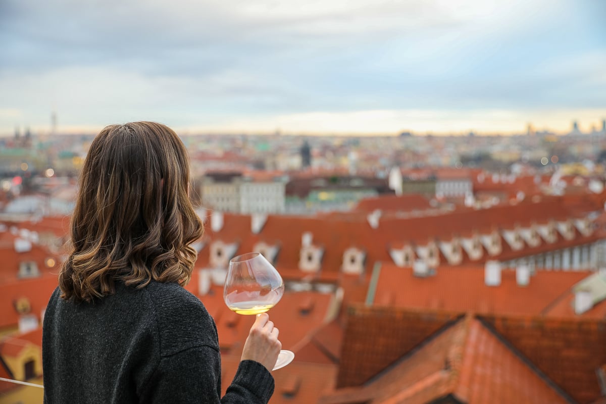 Maddy holding a glass of white wine while enjoying panoramic views of Prague’s red rooftops from Terasa U Zlaté Studně rooftop restaurant.