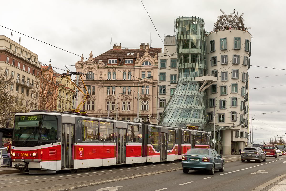 Red Prague tram passing in front of the Dancing House (Fred and Ginger building), with historic architecture and city traffic on a cloudy day.