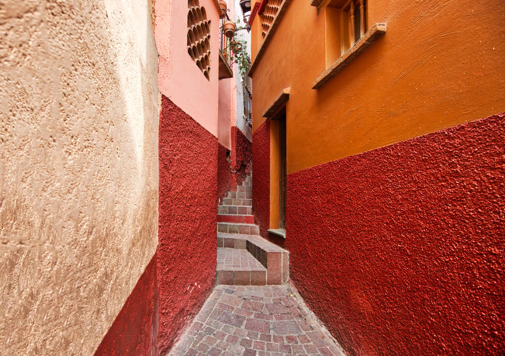 A narrow and winding alley in Mexico lined by pink, red, and orange buildings