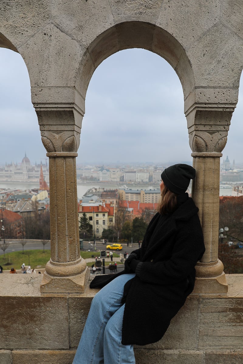 Maddy sitting between arched columns at the Fisherman's Bastion's lookout point overlooking Budapest