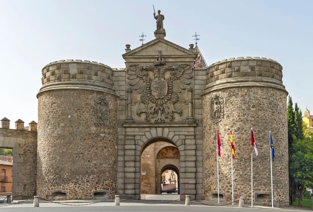 The Gate of Bisagra on a sunny day with 4 flags in front.