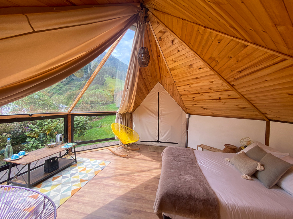 A big cozy bed with minimalist table and chairs inside a tent-cabin. The sunlight can easily pass through the roof-to-ceiling glass window making the room bright even without light bulbs.