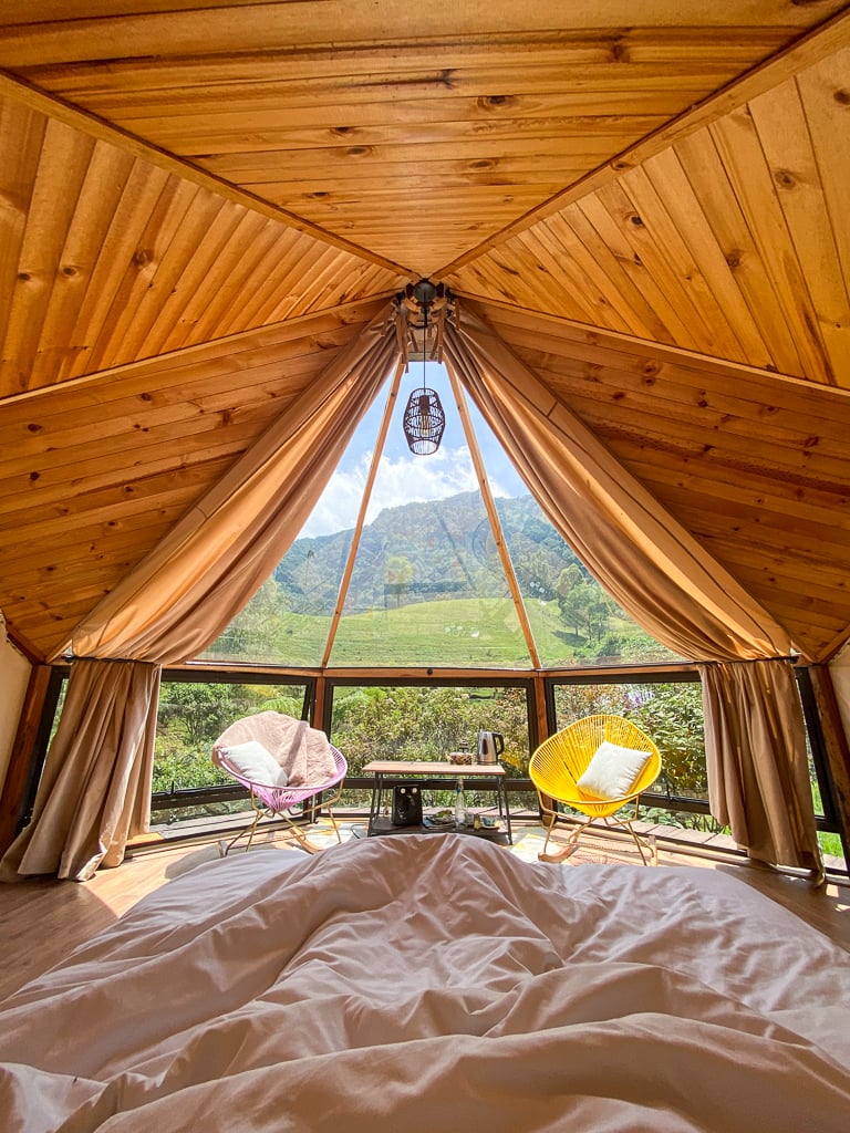 View of a tent-cabin from the inside with white beddings and cozy chairs. The floor-to-ceiling windows offer a magical view of the nature outside on a sunny day.