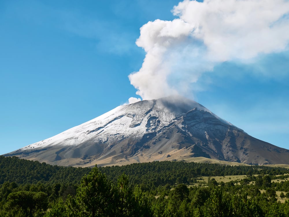 View of the Popo Volcano spewing smoke with green trees in the foreground