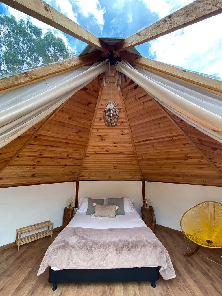 A warm cozy bed in the center of a tent-cabin under a roof divided in wood and glass. You can see the blue sky from the inside, which is one of the coolest experiences while glamping in Salento.