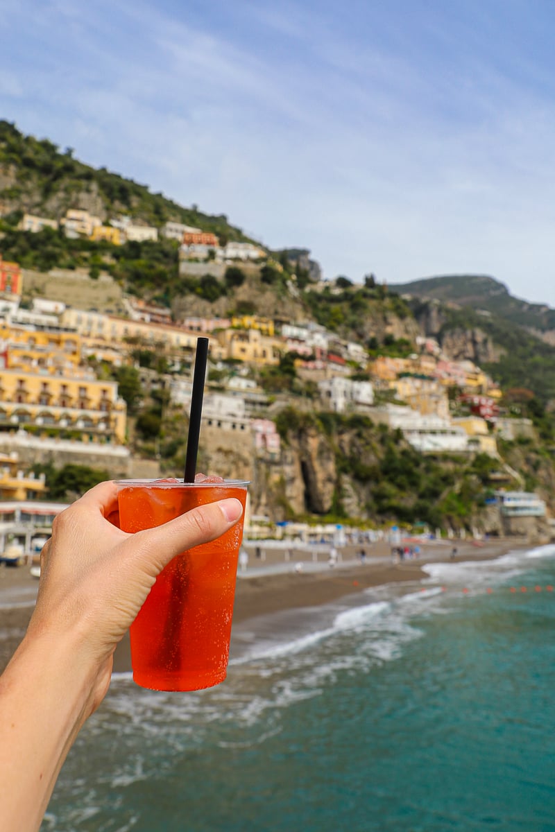 Maddy holding an Aperol spritz in a to-go cup with the town of Positano in the background