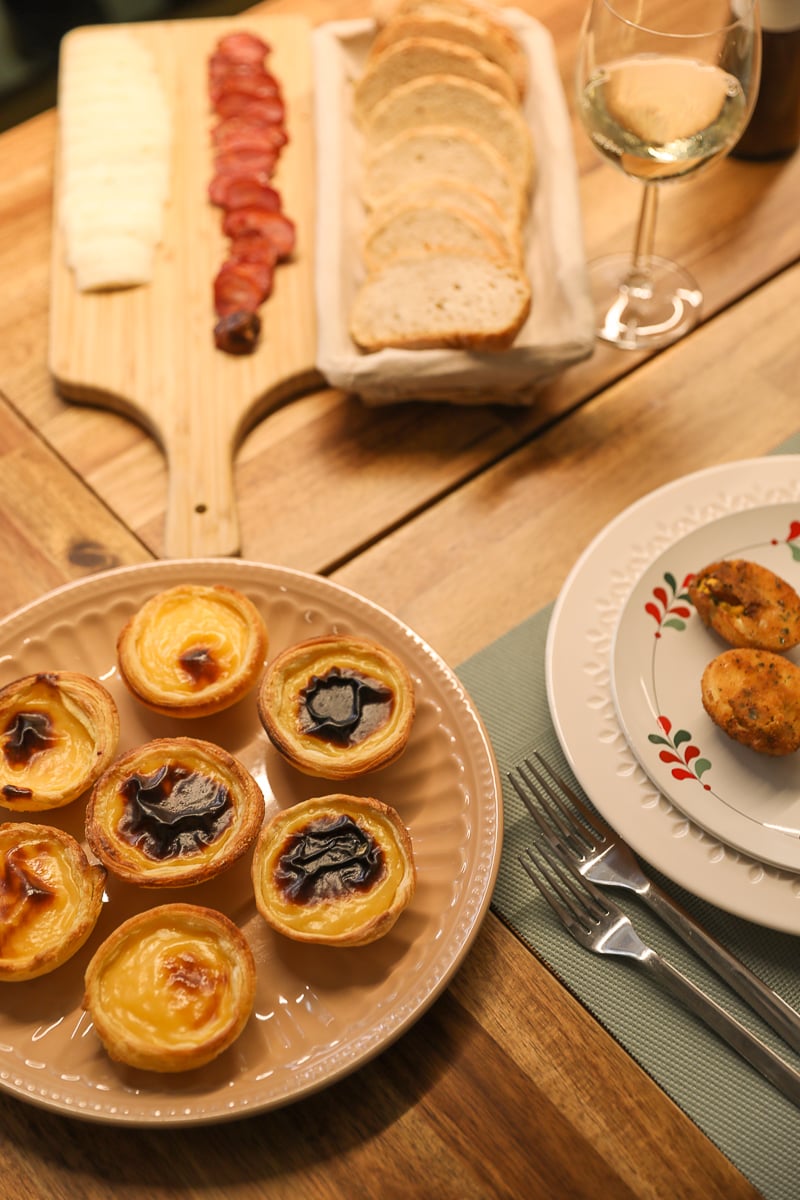 Freshly-baked pastel de natas, ovos verdes, slices of bread, and a glass of wine
