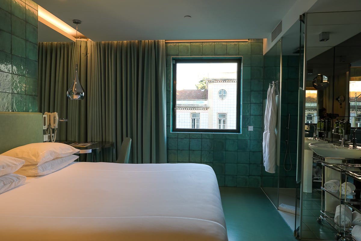 Bed against a tiled wall facing a glass-enclosed bathroom at WC Hotel