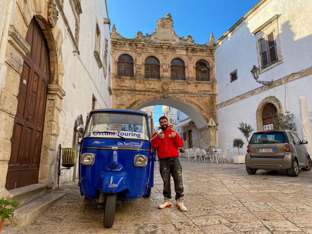 Man standing beside a blue tuk tuk near the Arco Scoppa. Taking a tuk tuk tour of the Millenary Olive Groves is one the best things to do in Ostuni.