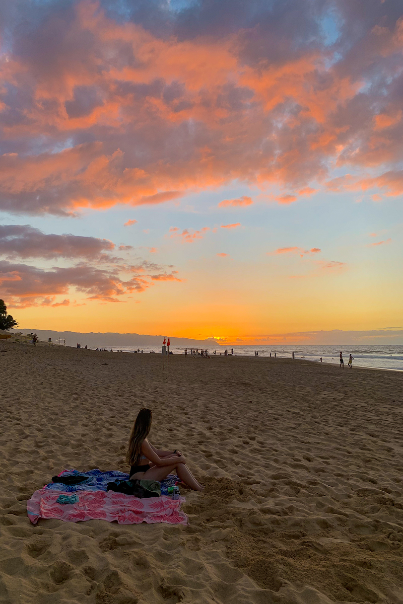 Maddy sitting on a sandy beach during sunset in the North Shore. In this Oahu itinerary, you'll find everything you need to know when visiting the island.