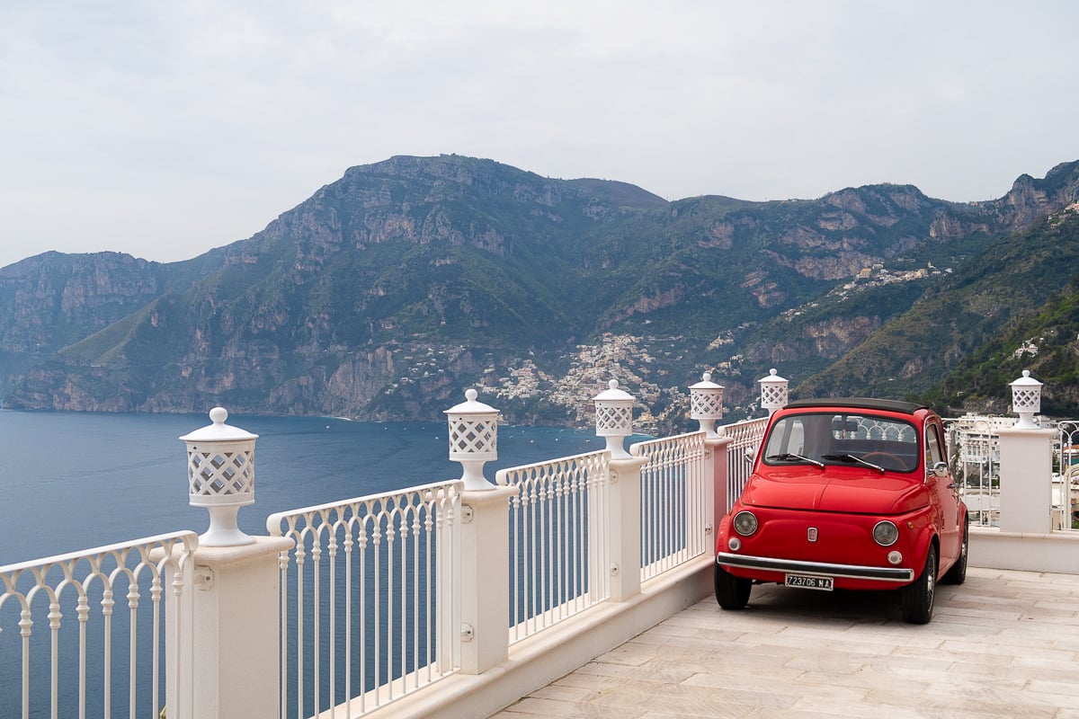 A vintage fiat parked on an ornate terrace overlooking the Amalfi Coast - this Amalfi Coast itinerary includes all of the must-know transportation details for a successful trip.