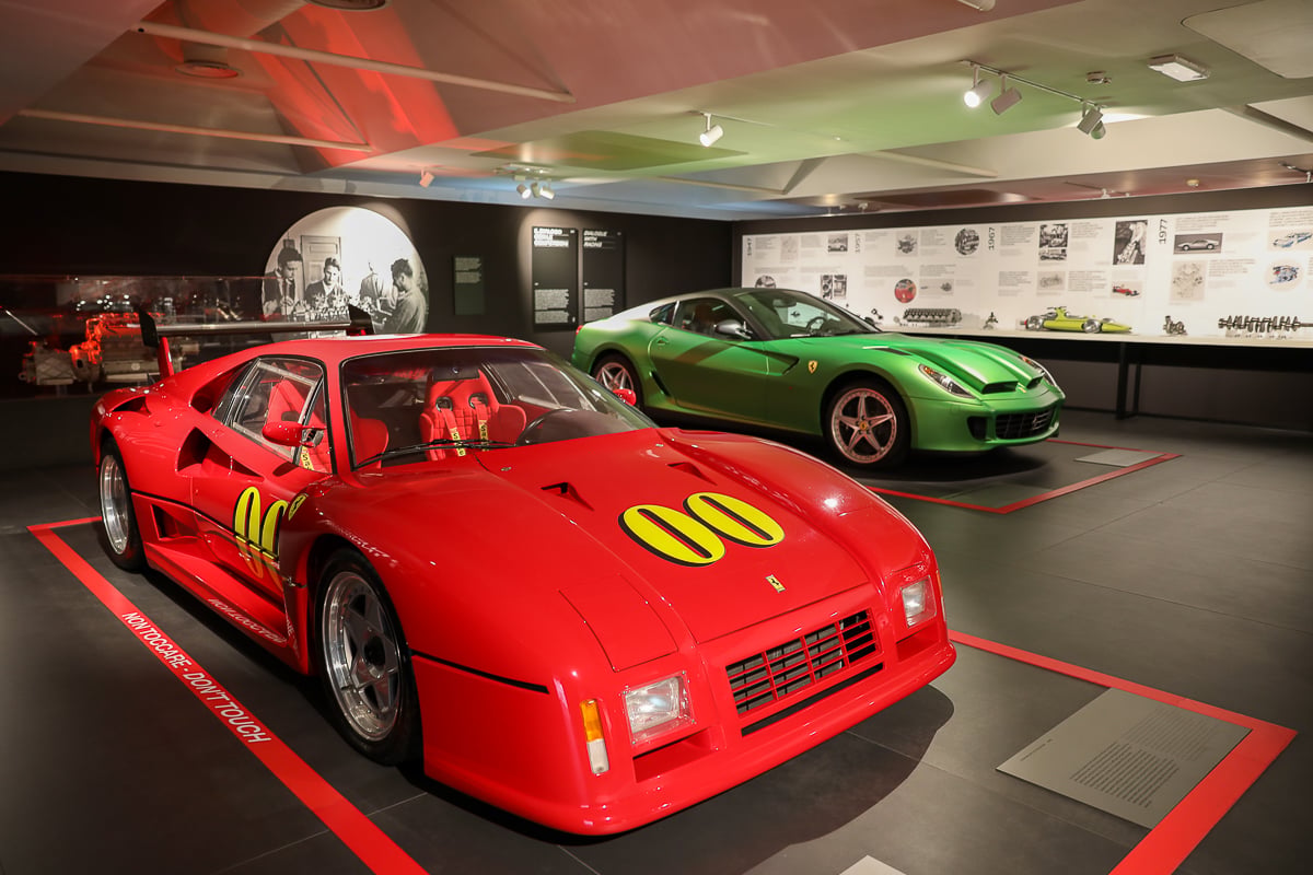 Red Ferrari race car with yellow number decals on display at the Maranello Ferrari Museum, with a green Ferrari sports car exhibited in the background.