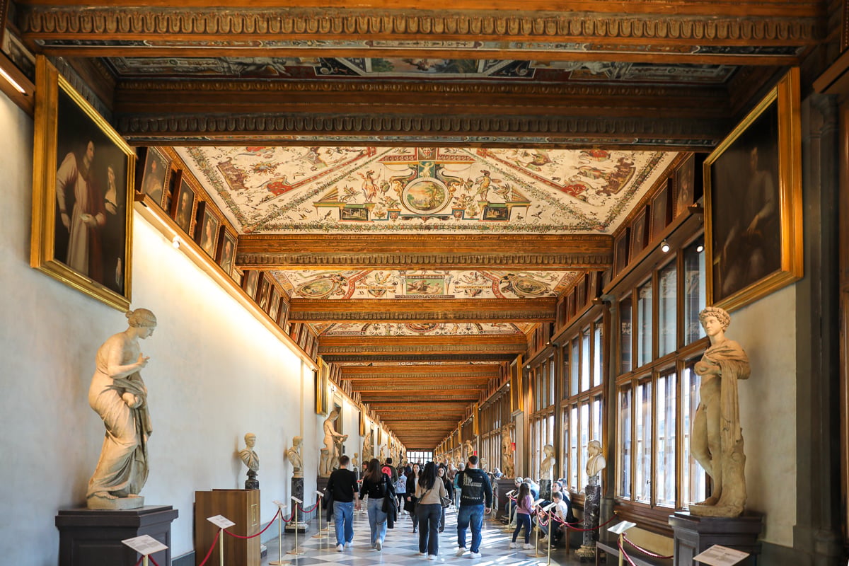An ornate hall filled with museum-goers and lined with statues within the Uffizi Gallery in Florence