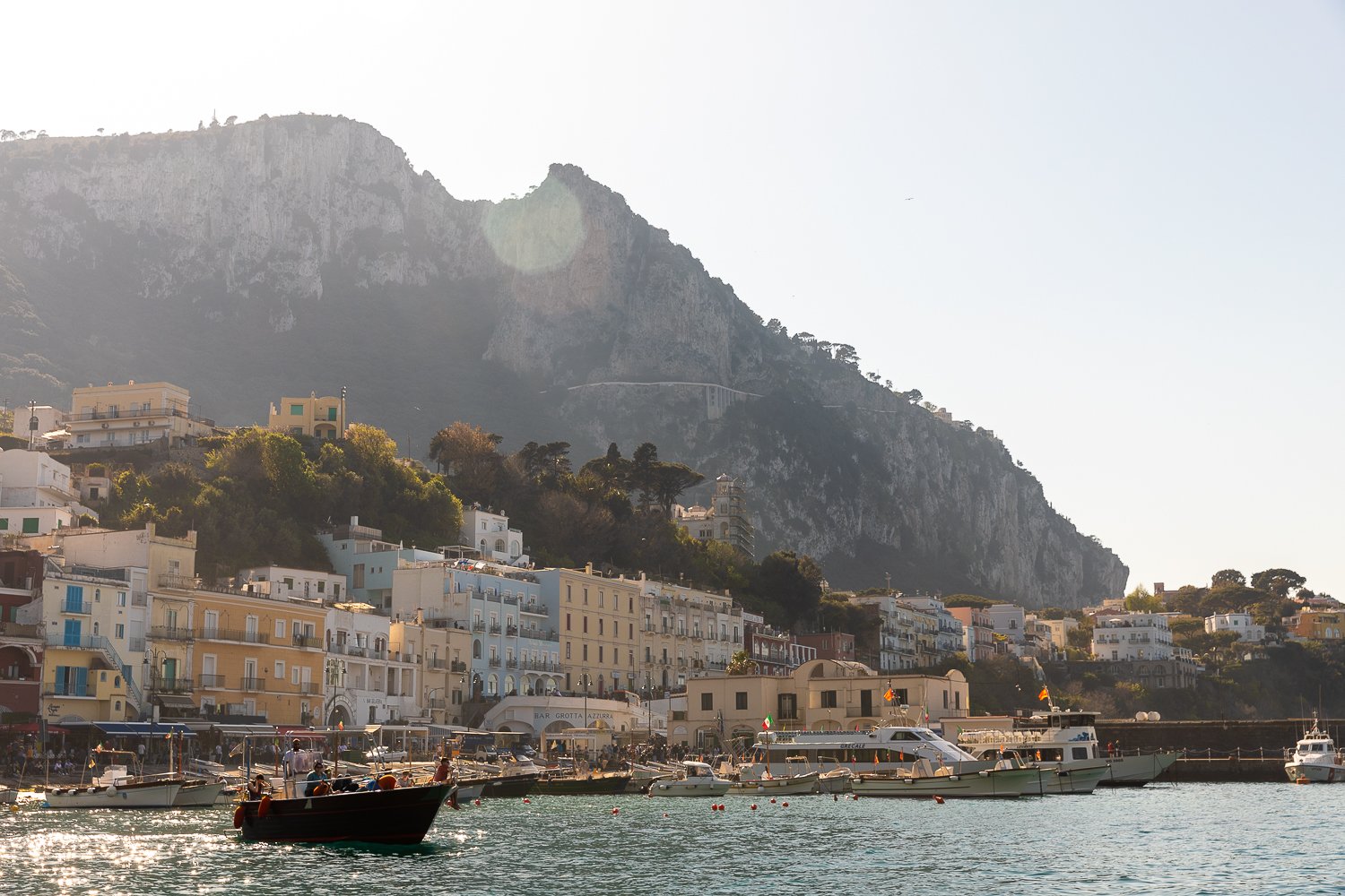 Group of people riding a boat with the view of Capri's colorful buildings and mountain on the background under the sunny sky