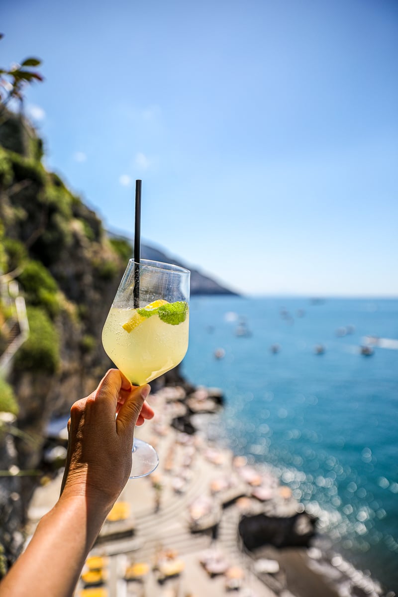 A hand holding a limoncello sproitz with sea views in the background