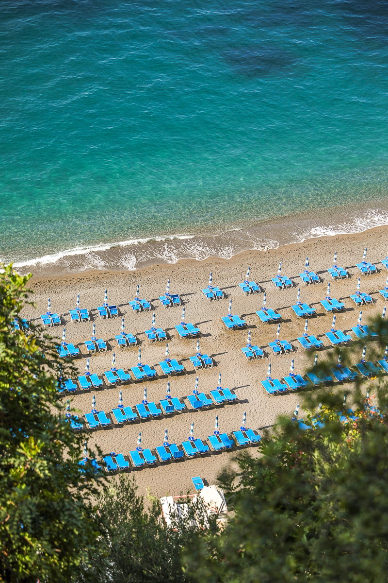 Neat rows of blue sunbeds and umbrellas on a Positano beach, viewed from above with turquoise water lapping the shore