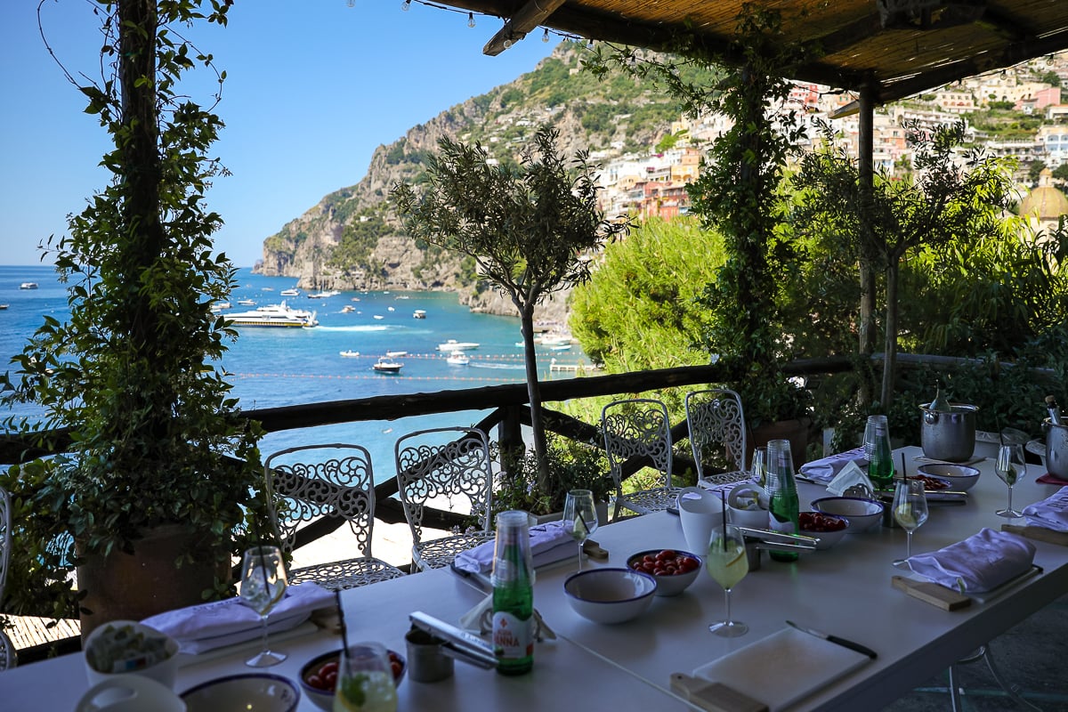 A white table with cutting boards, glasses, water bottles, and other cooking class items with a sea view in the background