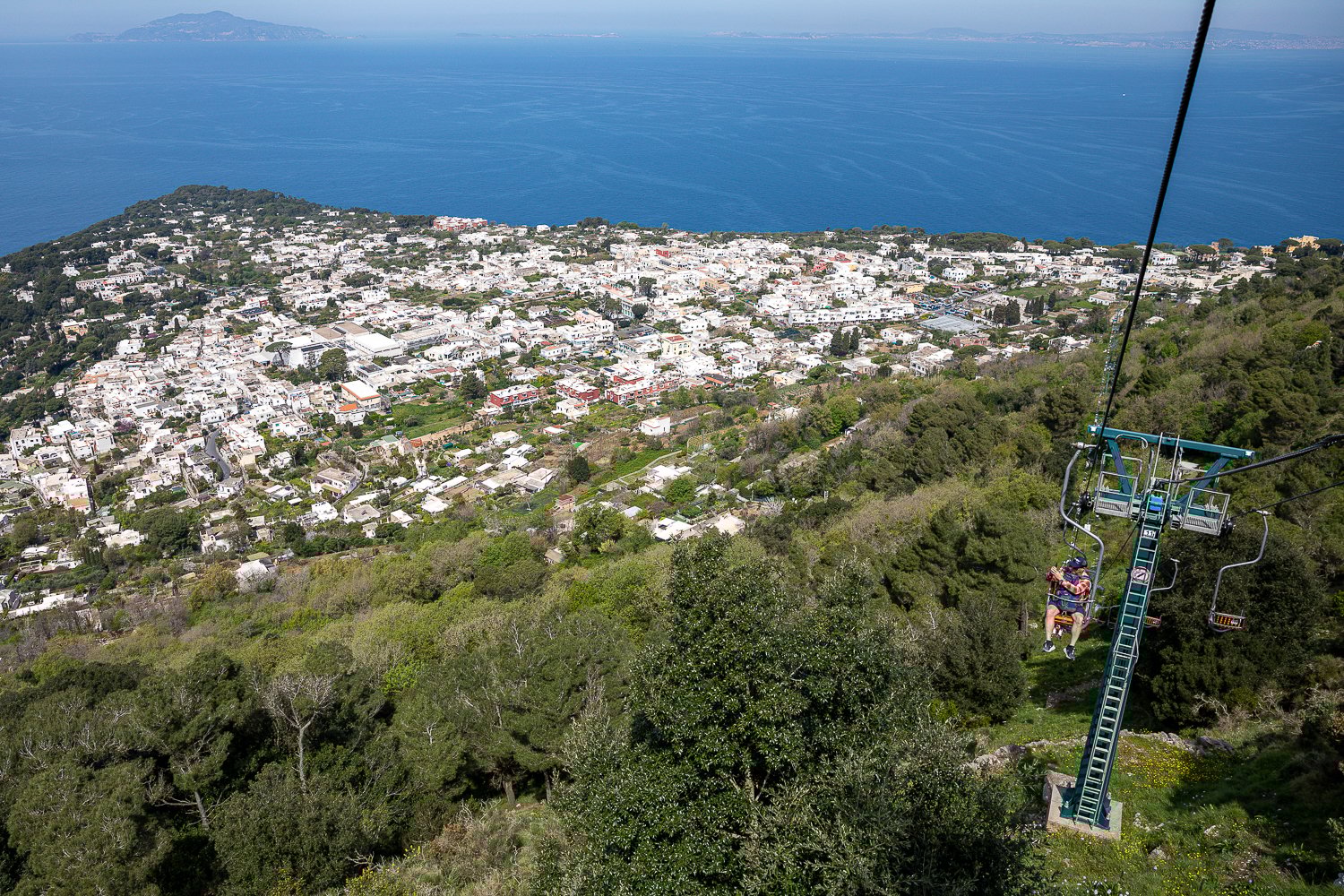 People riding the chairlift above the breathtaking views of Capri's lush greeneries and light-colored buildings as well as the blue sea