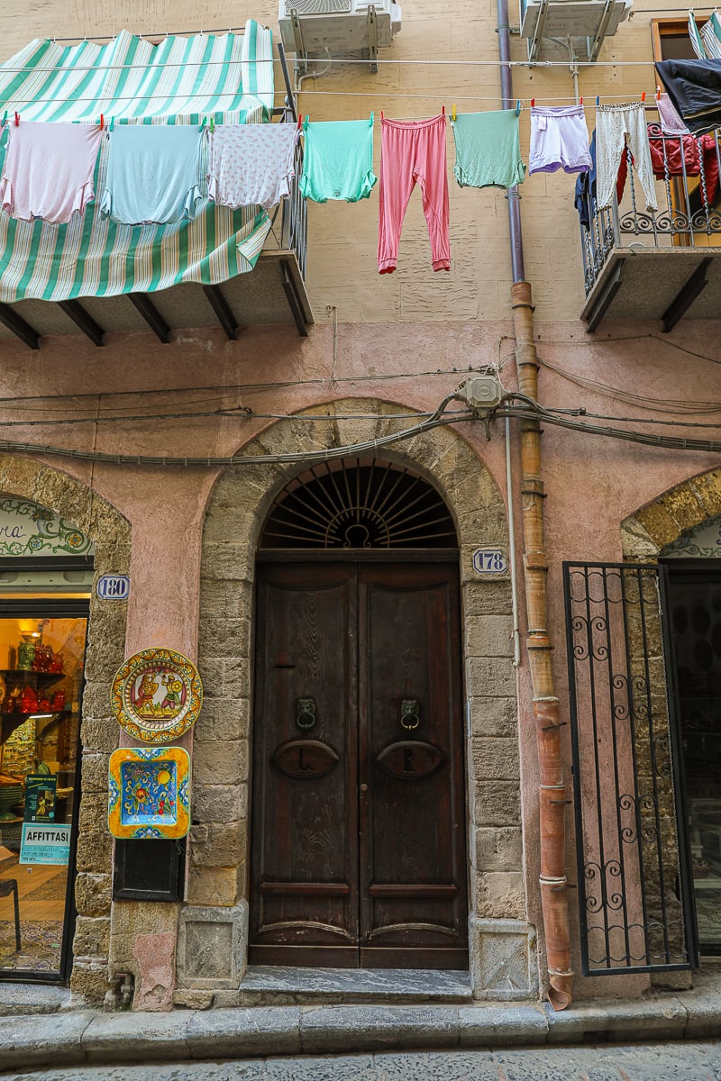 An arched wooden door along a street in Cefalù with colorful clothesline hanging from above
