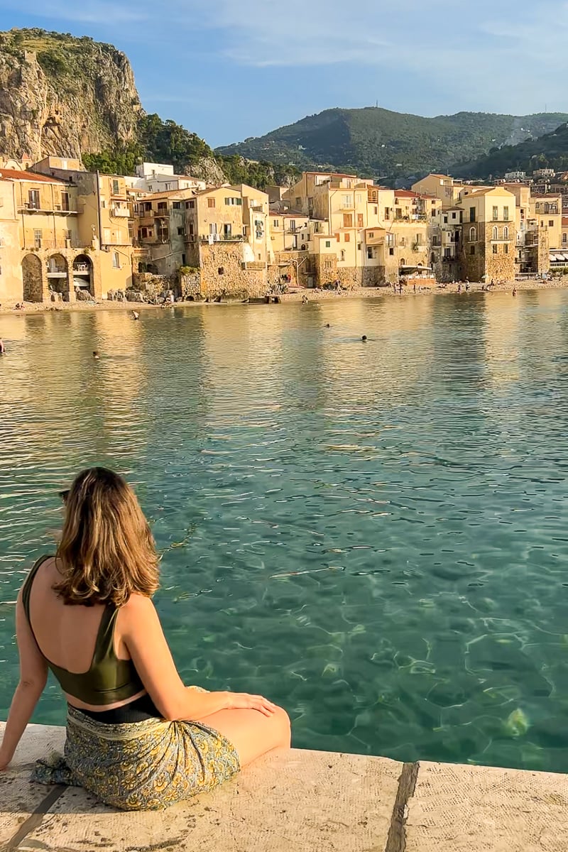 Maddy sitting by the sea while admiring the charming seaside city of Cefalu in Sicily