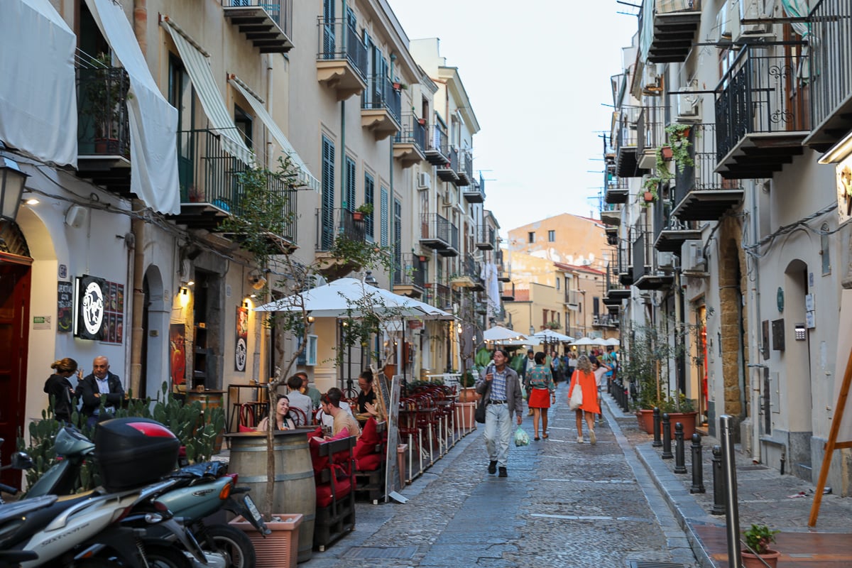People strolling along a narrow street in Cefalù lined with bars