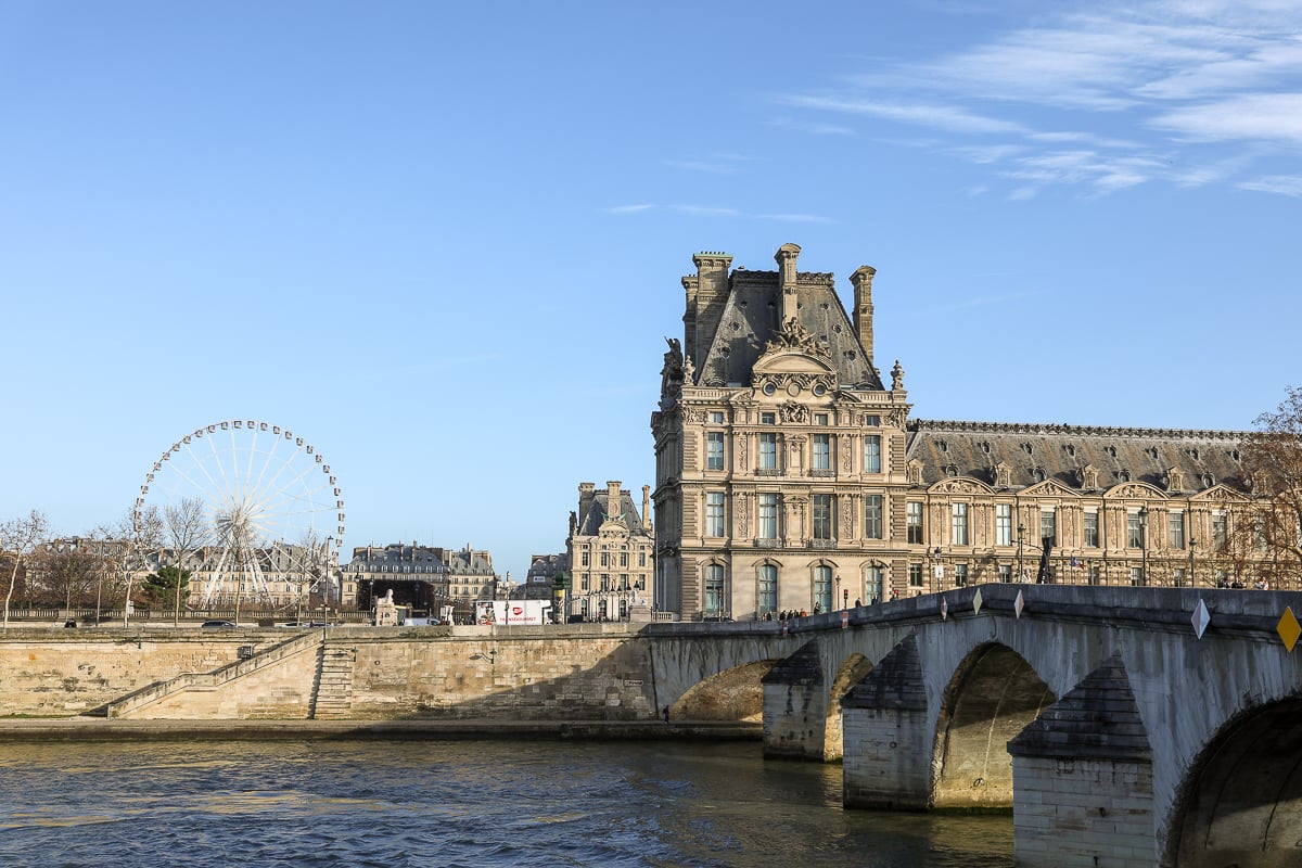 The Louvre Museum and the Ferris wheel in the Tuileries Garden, viewed from across the Seine River on a clear day.
