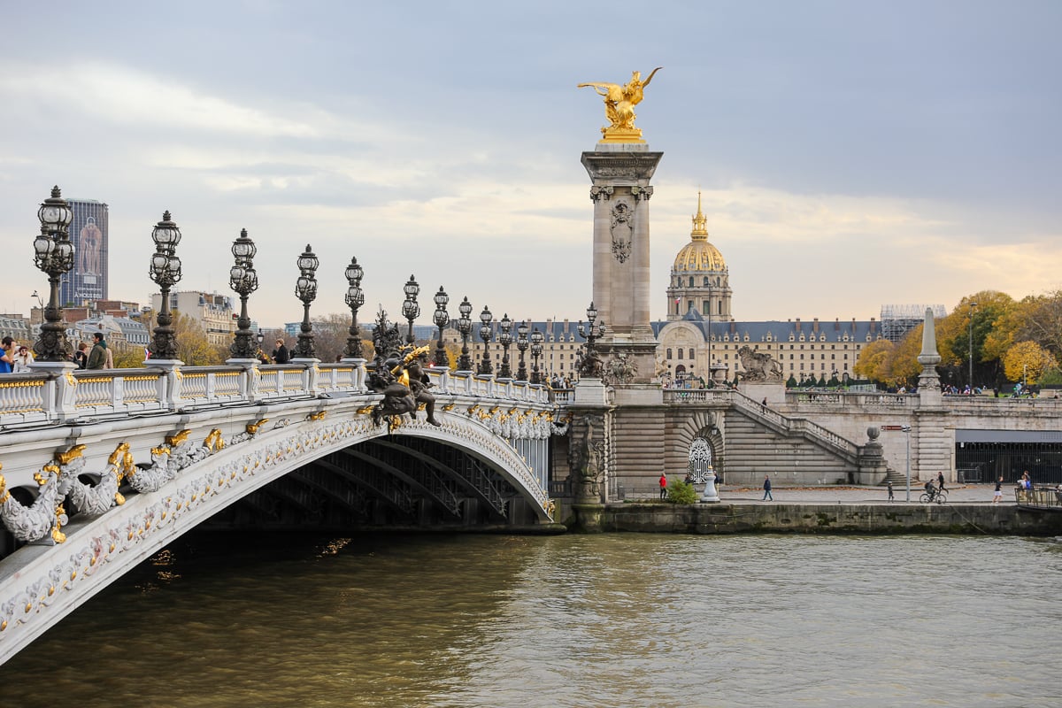 View of the ornate Pont Alexandre III bridge in Paris, with golden statues and the Hôtel des Invalides dome in the background.