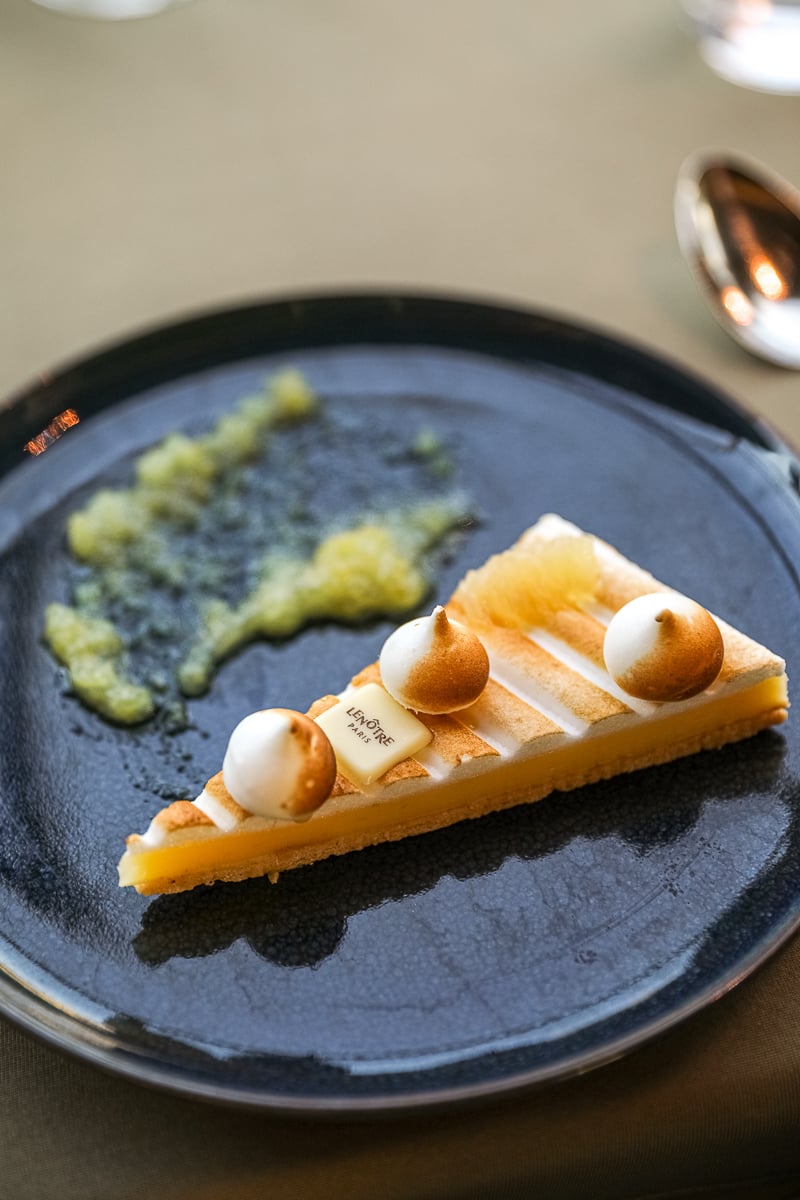 A slice of lemon tart from Lenôtre Paris, topped with toasted meringue and served with a fruit purée on a black ceramic plate.