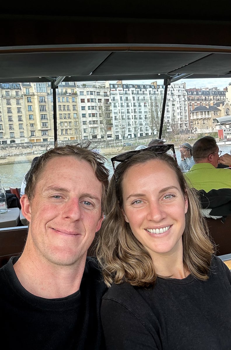 Maddy and Cacey, a couple, smiling on a covered river cruise boat along the Seine in Paris, with apartment buildings in the background.