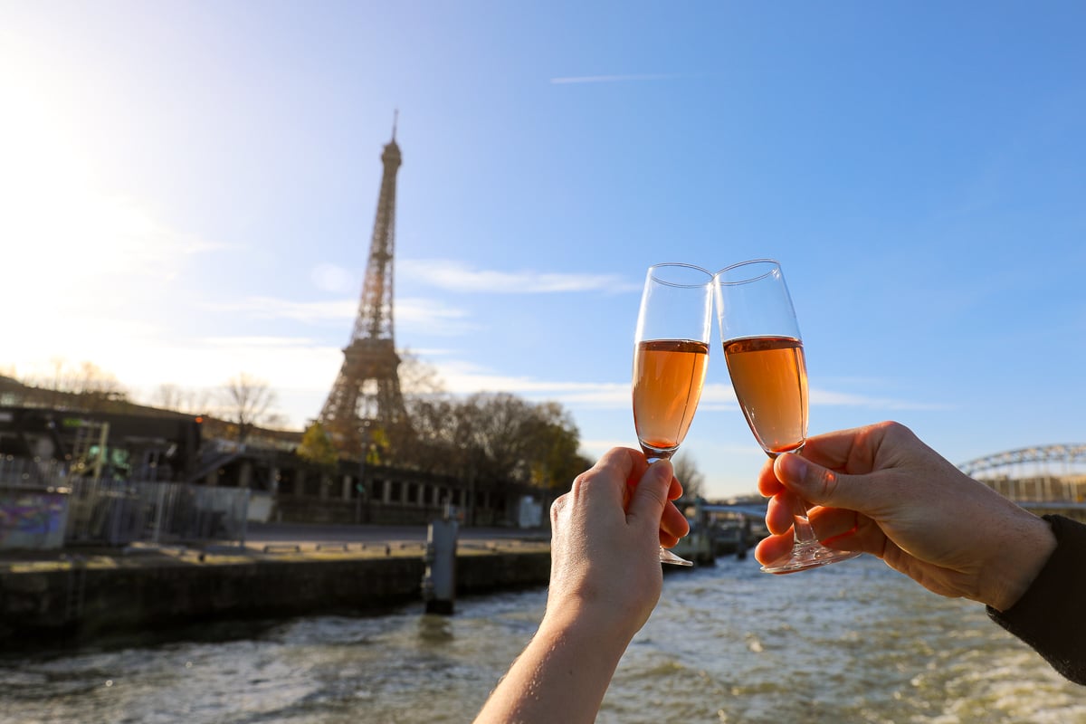Two people clinking glasses of rosé Champagne on a Seine River cruise in Paris, with the Eiffel Tower in the background on a sunny day.