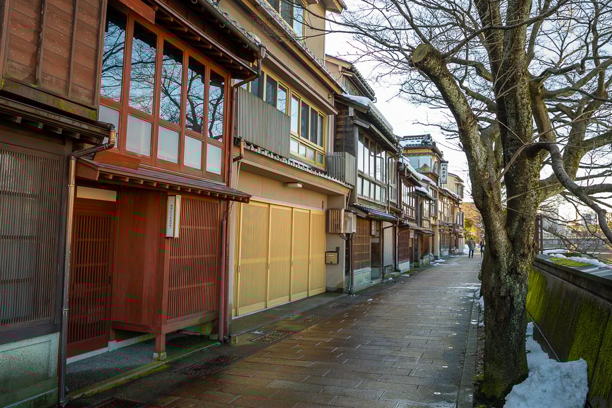 Quiet riverside street in Kanazawa bordered by traditional wooden machiya townhouses and moss-covered stone walls in winter.